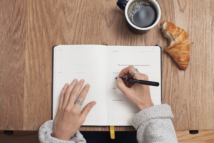 Mulher escrevendo em caderno sobre a mesa, com um copo de café e um croissant ao lado.